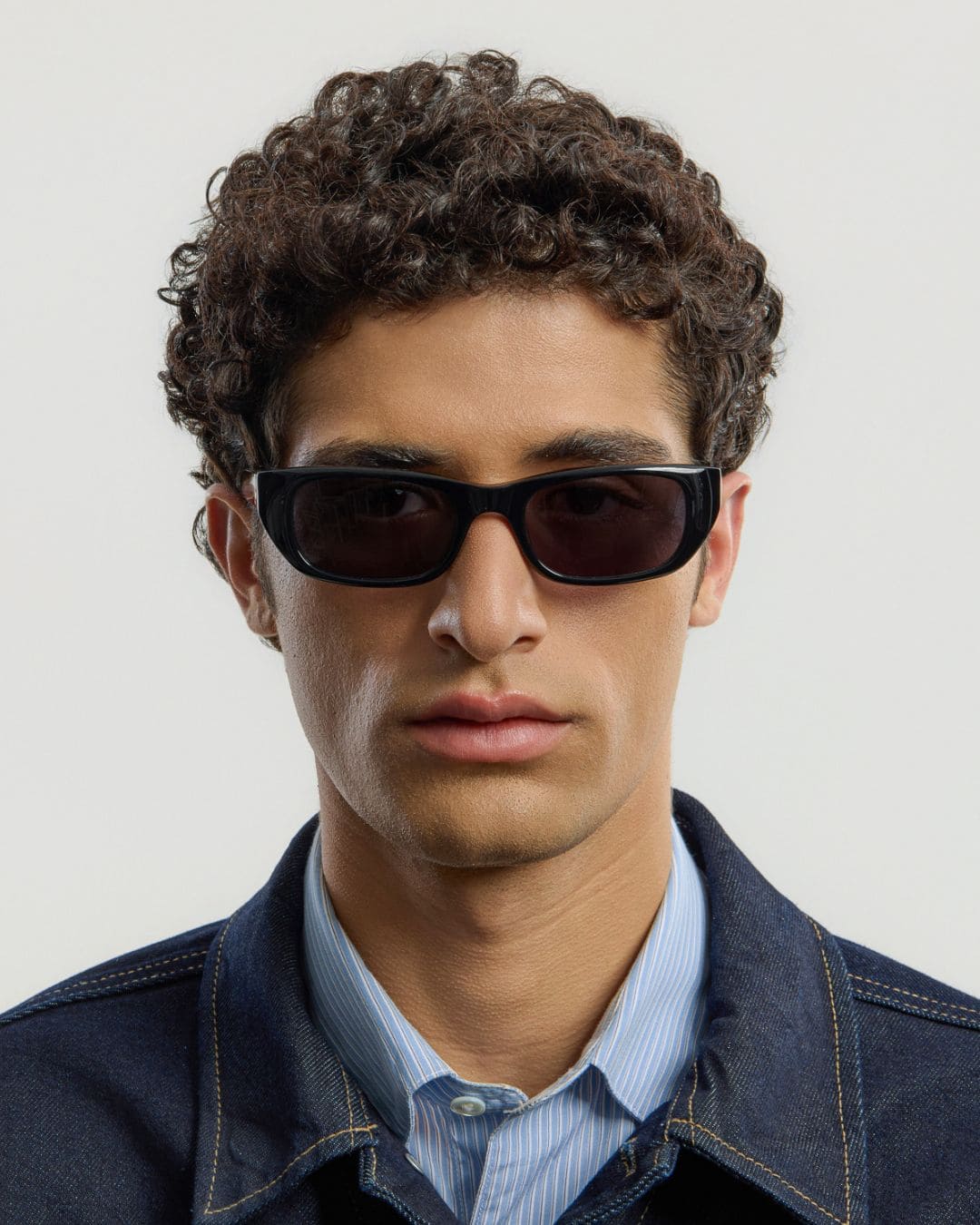 Man wearing black sunglasses and a denim jacket against a plain background